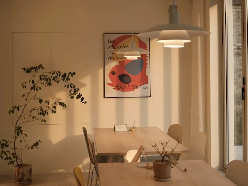 Warm ochre-walled room with a light timber table and chairs under a white pendant lamp, a tall leafy plant and a framed red and black poster