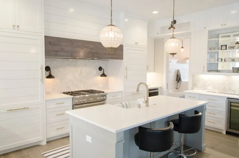 Bright white kitchen with a large island topped in white stone, black bar stools, a range cooker under a hood and two glass pendant lights