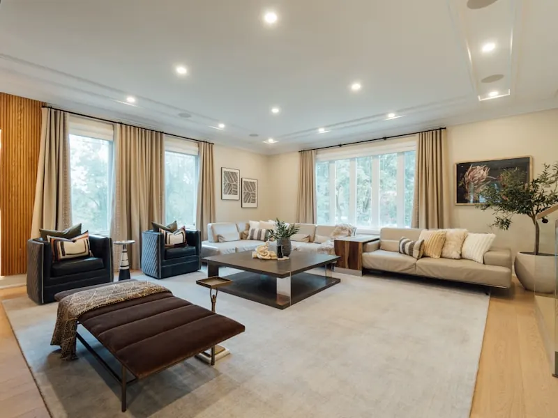 Spacious living room with recessed ceiling downlights, beige sofas around a low glass coffee table, a dark leather bench, a large rug and windows dressed in sheer curtains