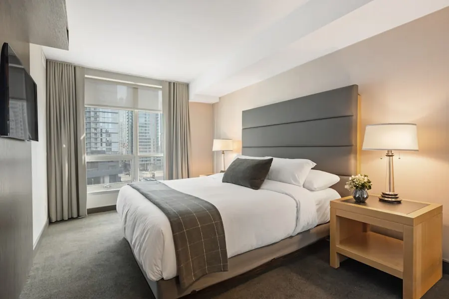 Bright bedroom with a padded grey headboard panel, crisp white bedding with a striped throw, a timber bedside table with a lamp and tall windows with grey curtains
